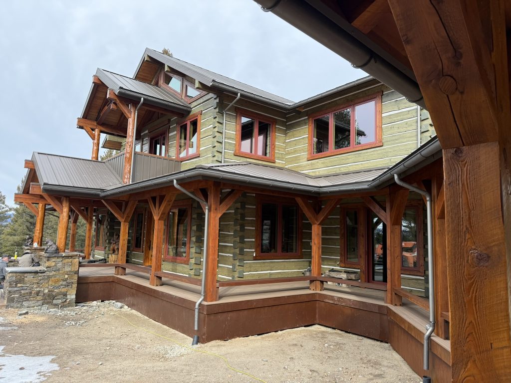 Large multi-story mountain home under construction with brown standing seam metal roof timber frame porch columns log and green siding red window trim and new gutters