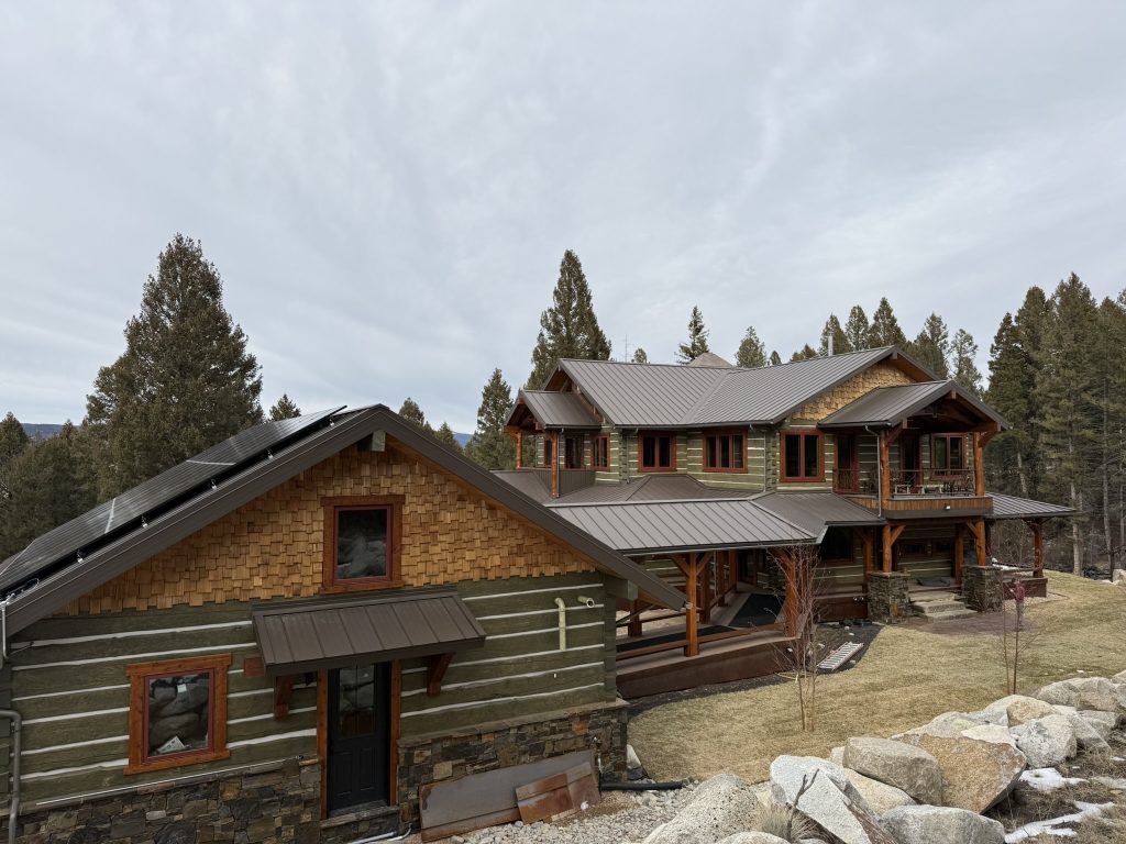 Wide view of large mountain home compound with brown standing seam metal roof solar panels on garage cedar shake gable green log siding stone base and timber frame porch