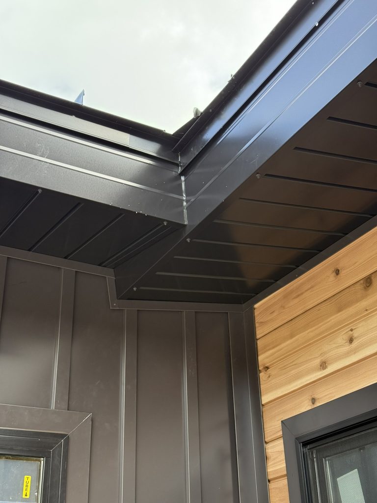 Close-up looking up at dark espresso metal soffit and fascia corner with cedar tongue-and-groove siding accent panel and board and batten metal wall panel meeting at eave