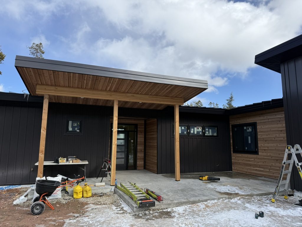 Modern new construction home entry with black board and batten metal siding natural cedar horizontal lap accent section and timber post flat-roof canopy with cedar soffit during construction