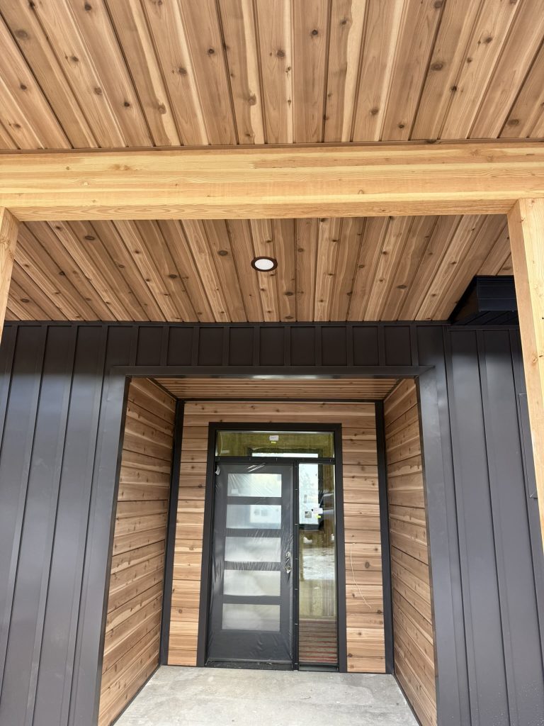 Interior view of covered entry porch with natural cedar tongue-and-groove soffit timber beam frame recessed light and black glass entry door surrounded by cedar horizontal siding