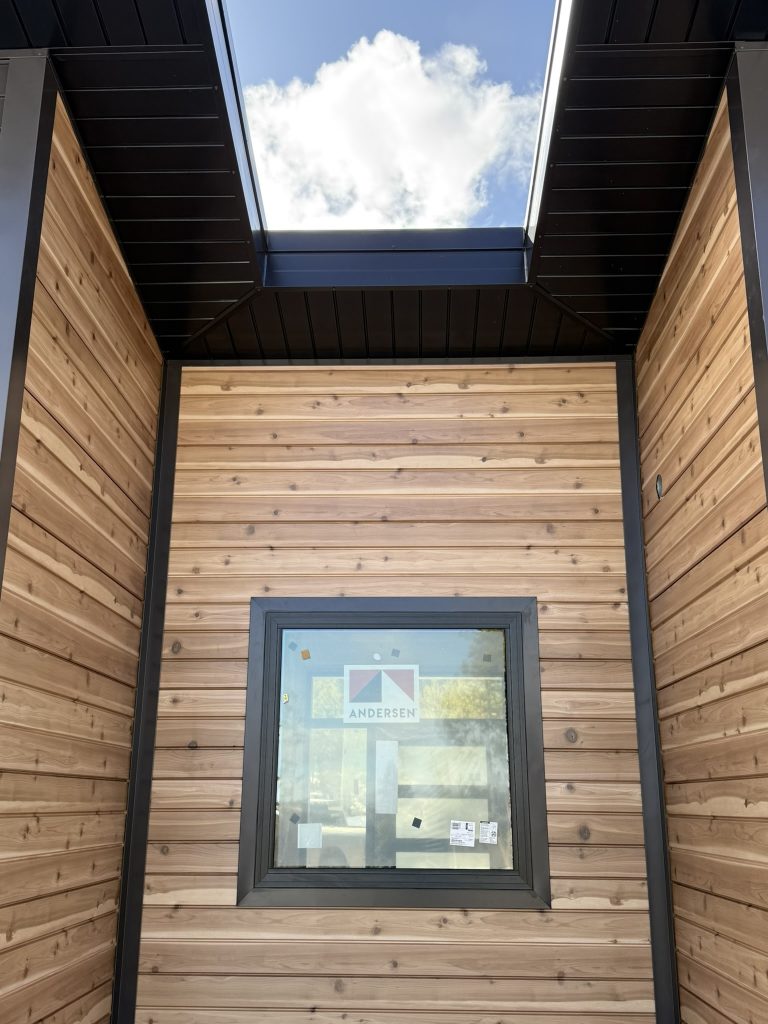 Looking up at cedar horizontal lap siding wall with black-framed Andersen window black metal soffit and board and batten panels from covered entry below blue sky