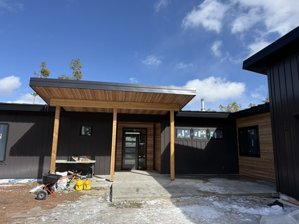 Front view of modern home entry with flat-roof standing seam canopy on timber posts cedar tongue-and-groove soffit black board and batten siding cedar horizontal lap accent and black glass door