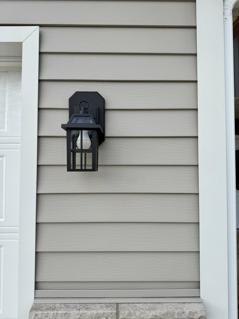 Close-up of black exterior wall lantern mounted on light gray horizontal lap siding between two white garage doors with dark charcoal board and batten upper siding
