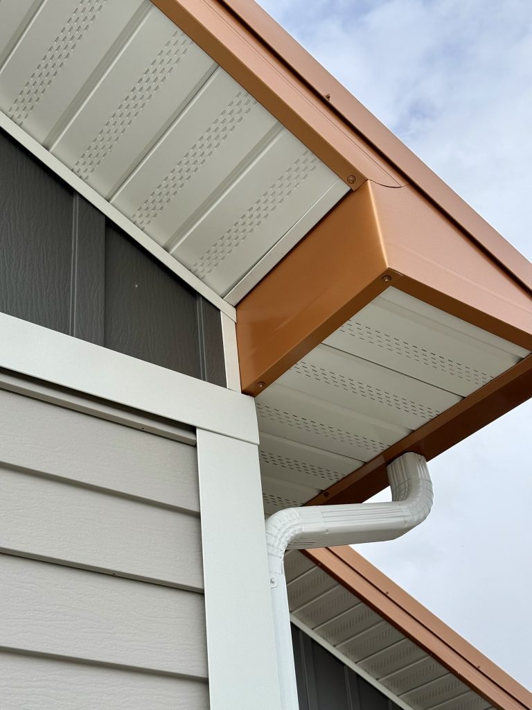 Close-up of copper penny-colored standing seam metal roof at eave with white vented soffit panels white downspout elbow and gray lap and board and batten siding