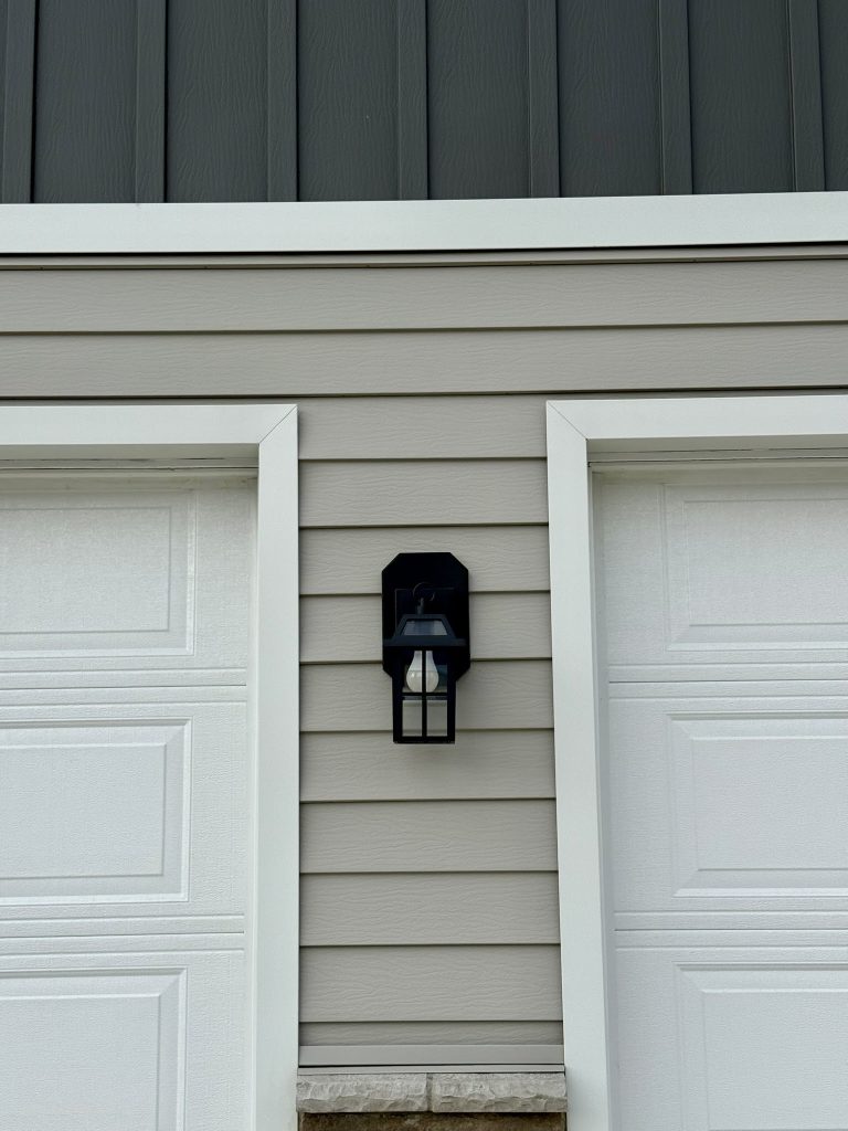 Black cage-style exterior wall lantern mounted between two white garage doors on gray horizontal lap siding with dark charcoal vertical board and batten upper gable