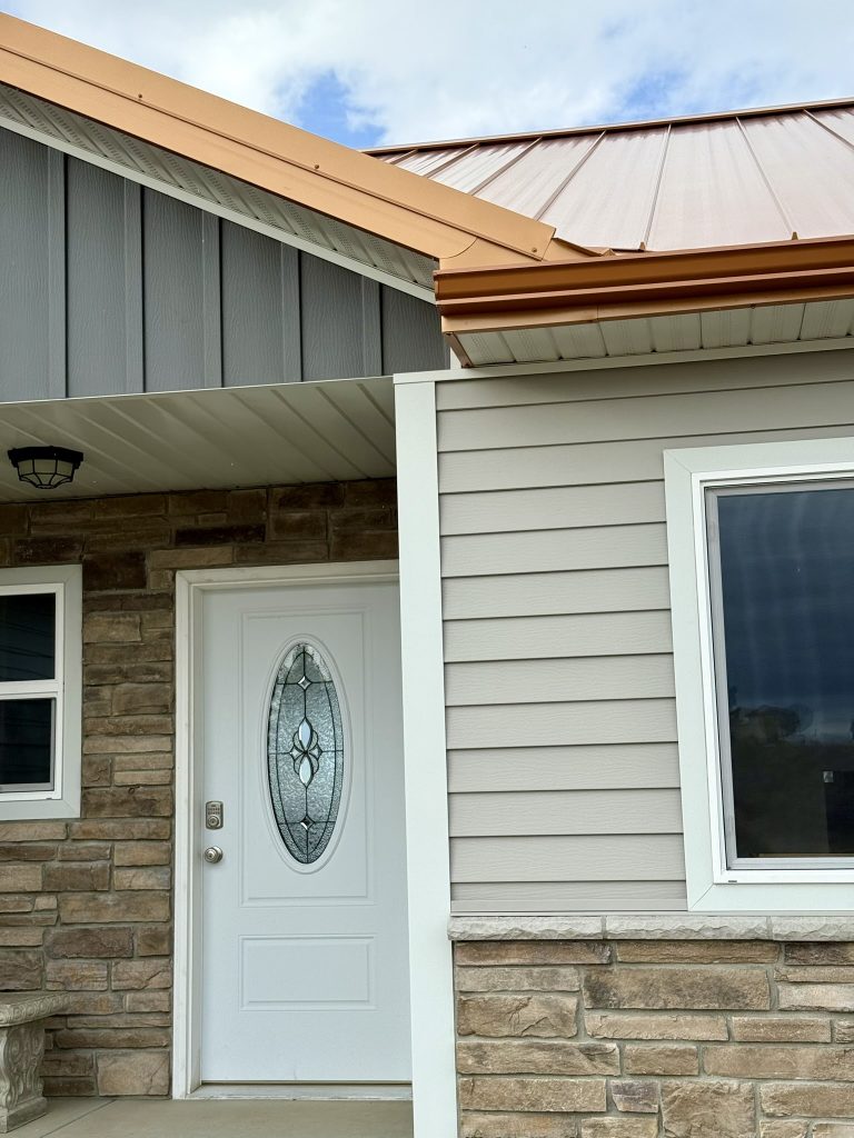 Completed entry porch with copper-tone standing seam metal roof gray horizontal lap siding white entry door with oval leaded glass insert and stone veneer base