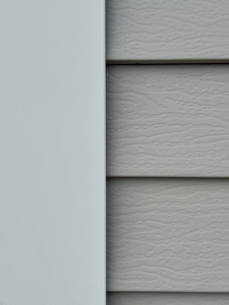 Close-up of white corner trim J-channel alongside light gray embossed lap siding panels showing panel overlap and corner installation detail