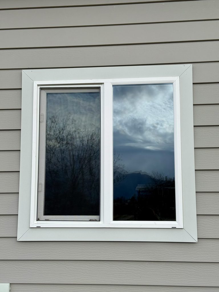 Close-up of newly installed double-pane window with white trim surround set into gray horizontal lap siding with mountain and sky reflection in glass