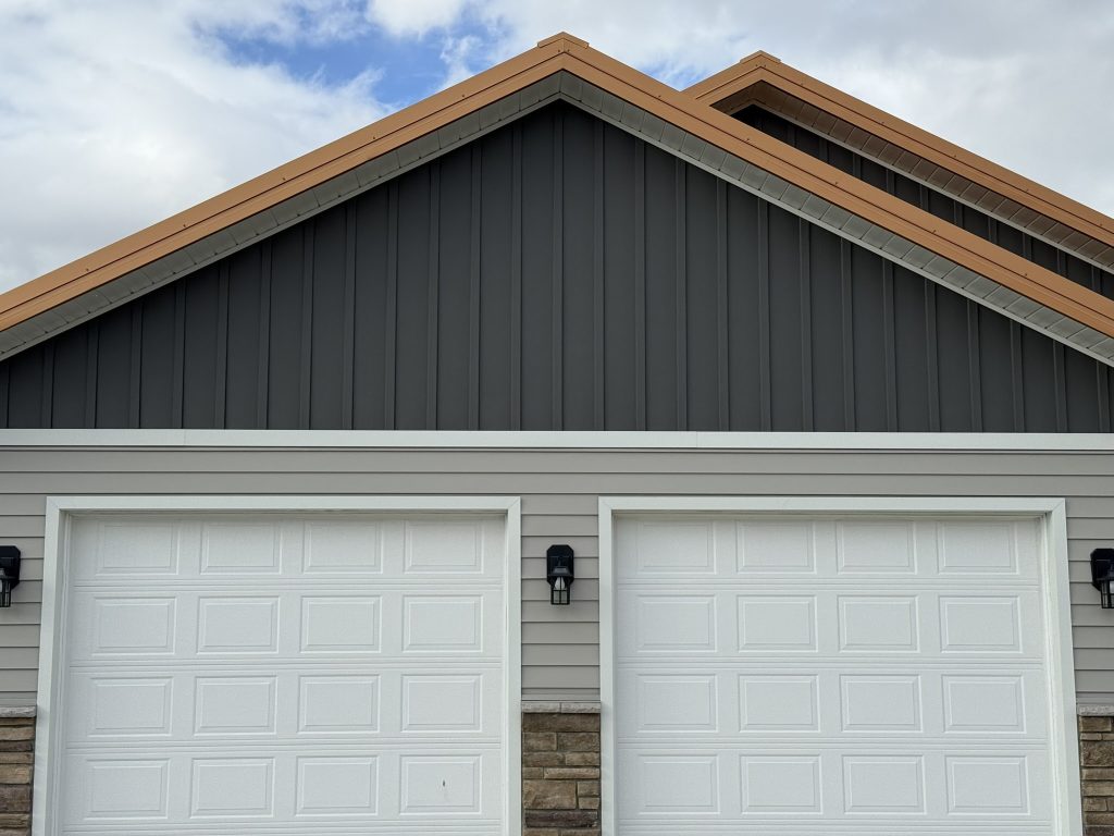 Garage gable end featuring dark charcoal vertical board and batten metal siding with copper-tone fascia trim above light gray horizontal lap siding and two white garage doors with stone base