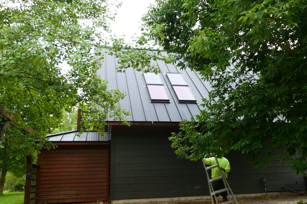 Crew member on ladder installing dark charcoal standing seam metal roof with flat skylight panels on a home with wood garage door surrounded by summer foliage