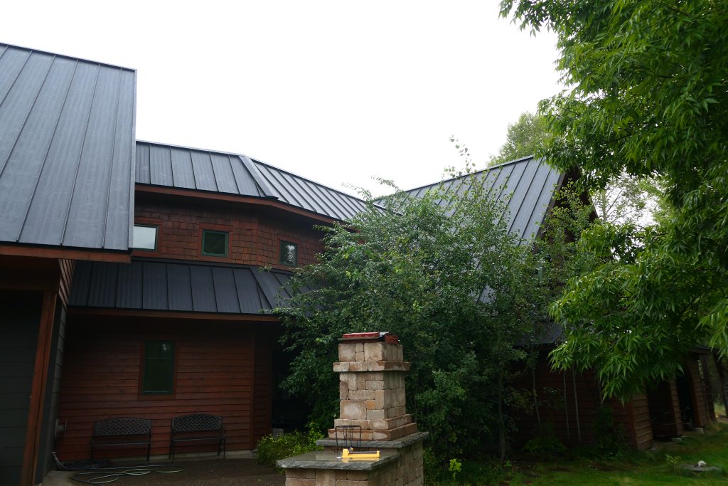 Rear view of large home with complex multi-plane dark charcoal standing seam metal roof over cedar shake siding green-trimmed windows and stone chimney surrounded by summer trees
