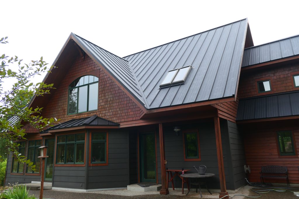 Front view of home with dark charcoal standing seam metal roof multiple gables integrated skylights cedar shake siding and covered entry porch with green-trimmed windows