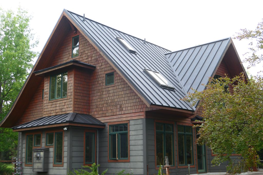 Completed dark charcoal standing seam metal roof with multiple skylights on craftsman home with cedar shake and gray lap siding green-trimmed windows and covered entry in summer