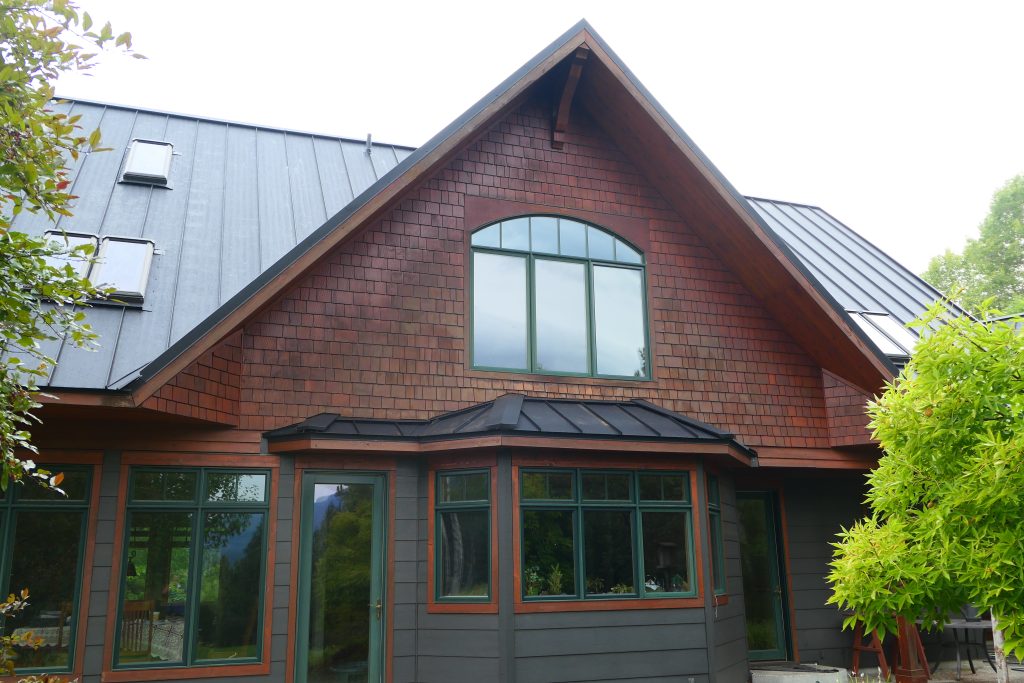 Craftsman home gable showing cedar shake siding arched window with green trim dark covered entry bump-out and dark charcoal standing seam main roof with skylights in summer foliage