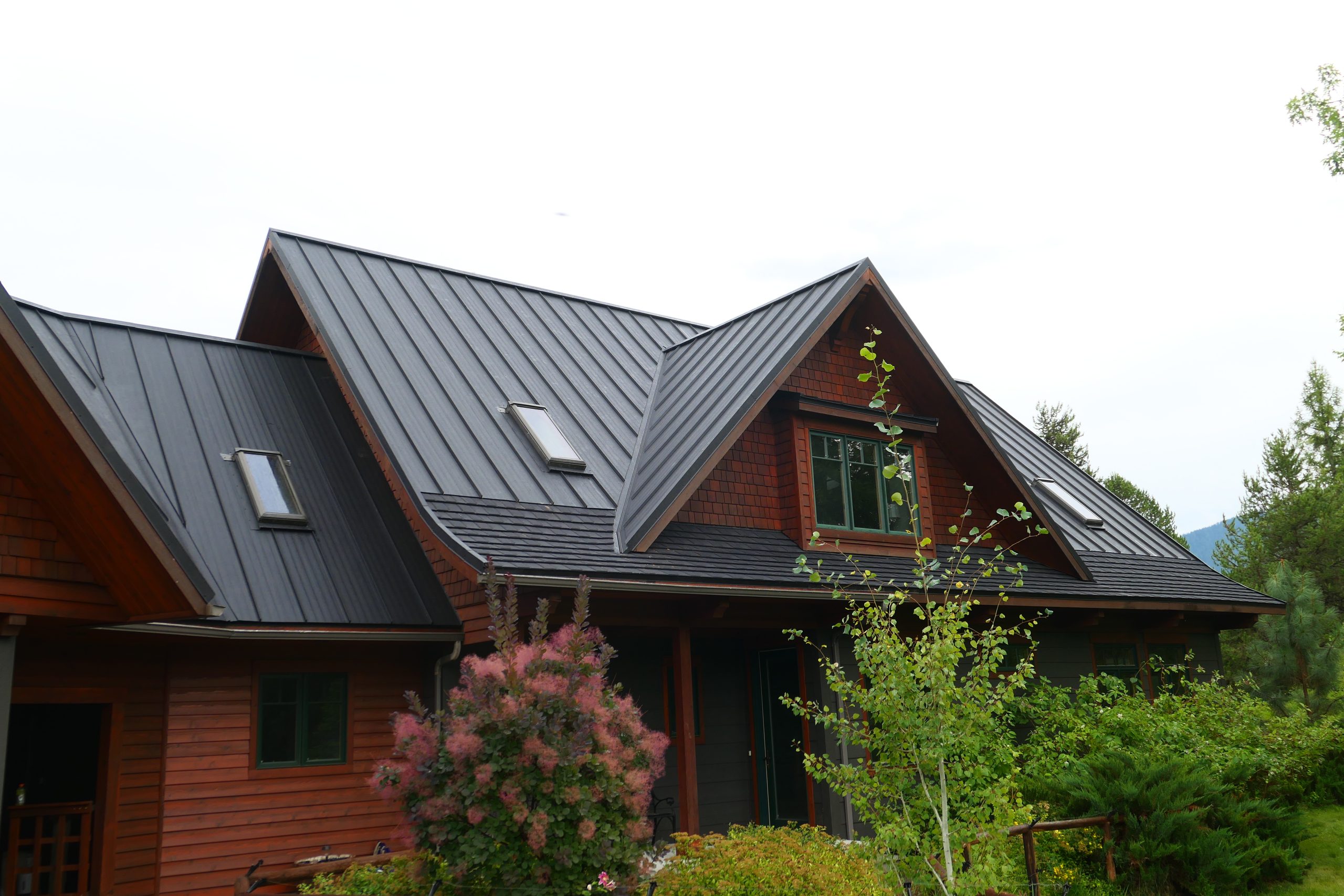 Full exterior of craftsman home with complex dark standing seam roof multiple skylights cedar shake upper siding gray horizontal lap lower siding green-trimmed windows and mature summer landscaping