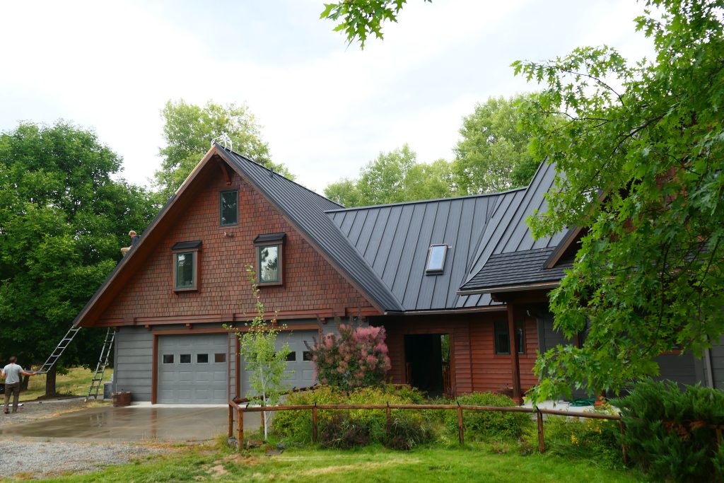 Standing seam metal roof installation in progress on craftsman home with crew member visible on roof cedar shake siding and ladder at garage in summer setting