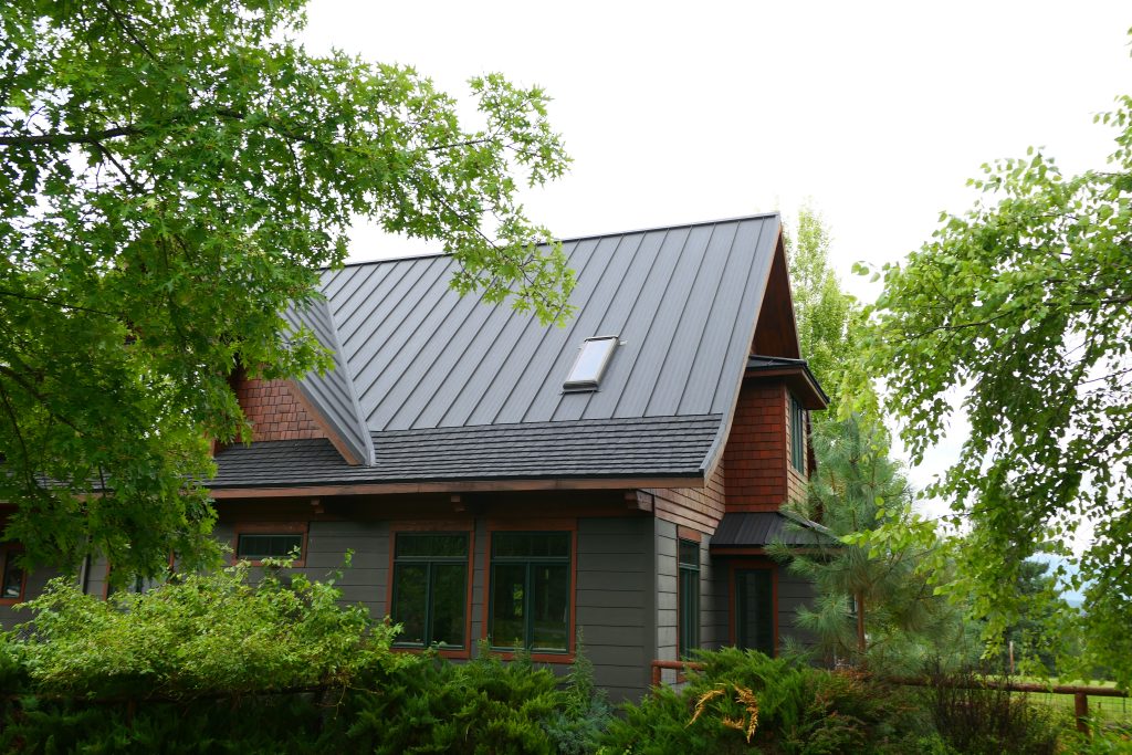 Side and rear view of craftsman home with completed dark standing seam roof with skylight cedar shake upper siding gray horizontal lap lower siding and mature summer landscaping