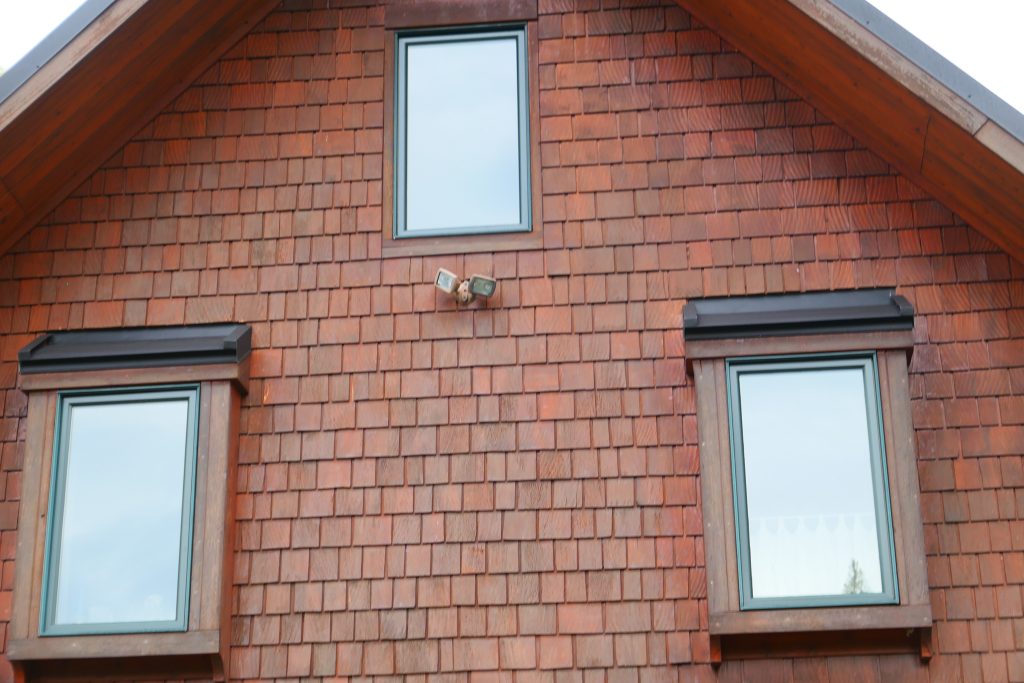 Close-up of cedar shake gable siding with dark metal window hood trim accents and green-trimmed windows beneath dark standing seam metal roof eave