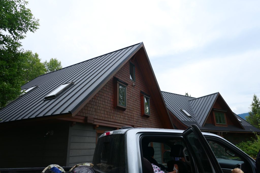Completed dark charcoal standing seam metal roof with integrated skylights on a multi-gable craftsman home with cedar shake siding and green-trimmed windows in summer foliage