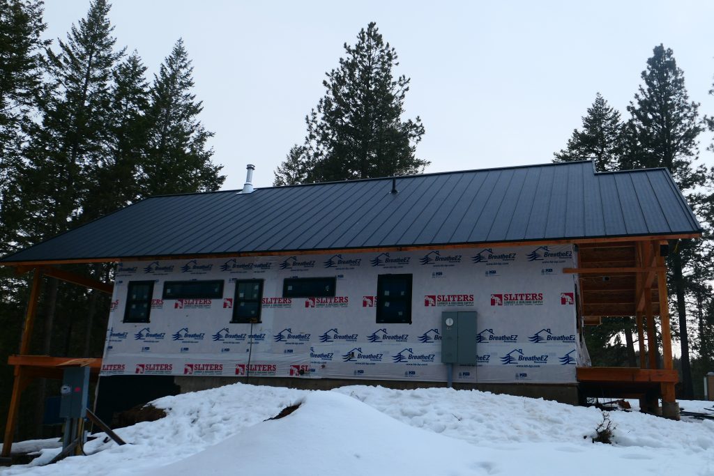 New construction home with completed dark standing seam metal roof still wrapped in BreatheEZ housewrap with timber frame porch structure and multiple black-framed windows in winter