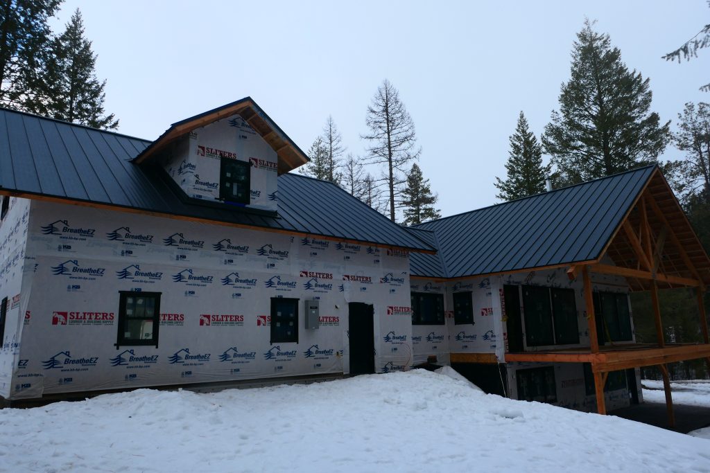 Side view of new construction home with dark standing seam metal roof timber frame covered porch structure and house wrap walls awaiting exterior siding in winter