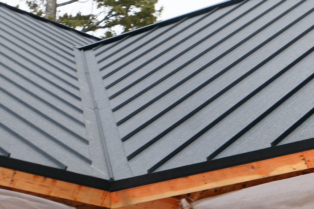 Close-up of freshly installed dark charcoal standing seam metal roof panel hip corner with exposed raw timber rafter tails and black eave trim showing panel layout and ridge detail
