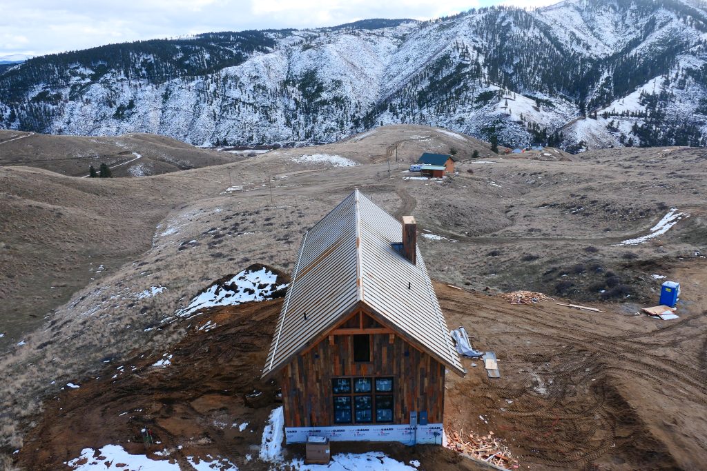 Aerial drone view of remote mountain cabin under construction with silver galvalume standing seam metal roof reclaimed barn wood vertical siding and dramatic snow-covered mountain canyon backdrop