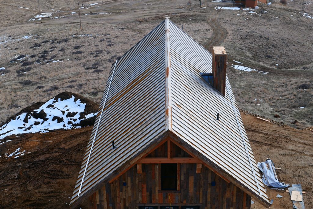 Aerial close-up of galvalume standing seam metal roof showing ridge panel layout chimney framing and reclaimed wood siding gable on remote mountain hillside construction site
