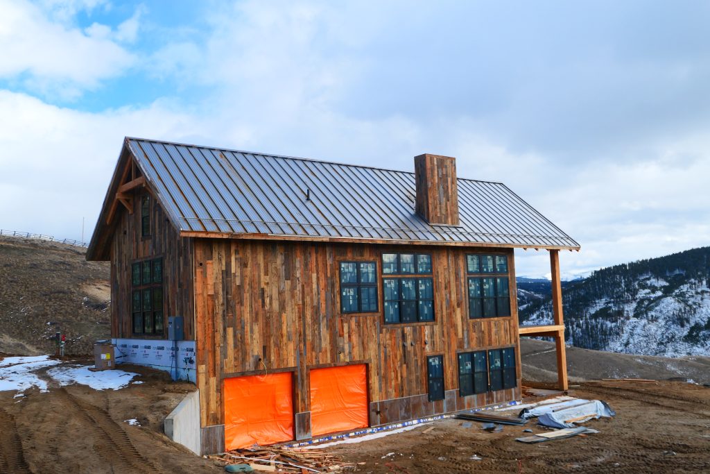 Ground-level view of remote mountain cabin with silver galvalume standing seam roof reclaimed barn wood siding timber frame gable accent large black-framed windows and orange tarps covering opening