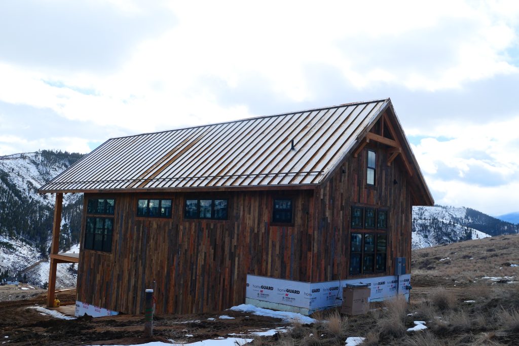 Front view of remote mountain cabin with silver galvalume standing seam roof reclaimed wood vertical siding multiple large black-framed windows and snowy mountain backdrop