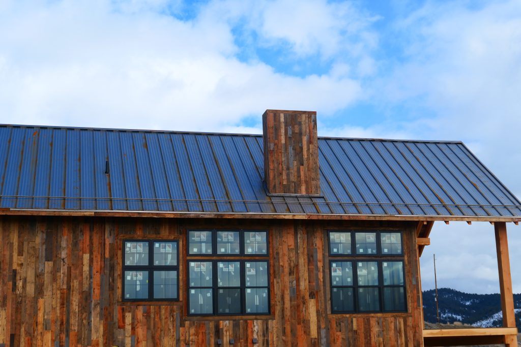 Close view of remote mountain cabin roofline with silver galvalume standing seam panels reclaimed barn wood chimney surround and large grid-pattern black-framed windows against open sky